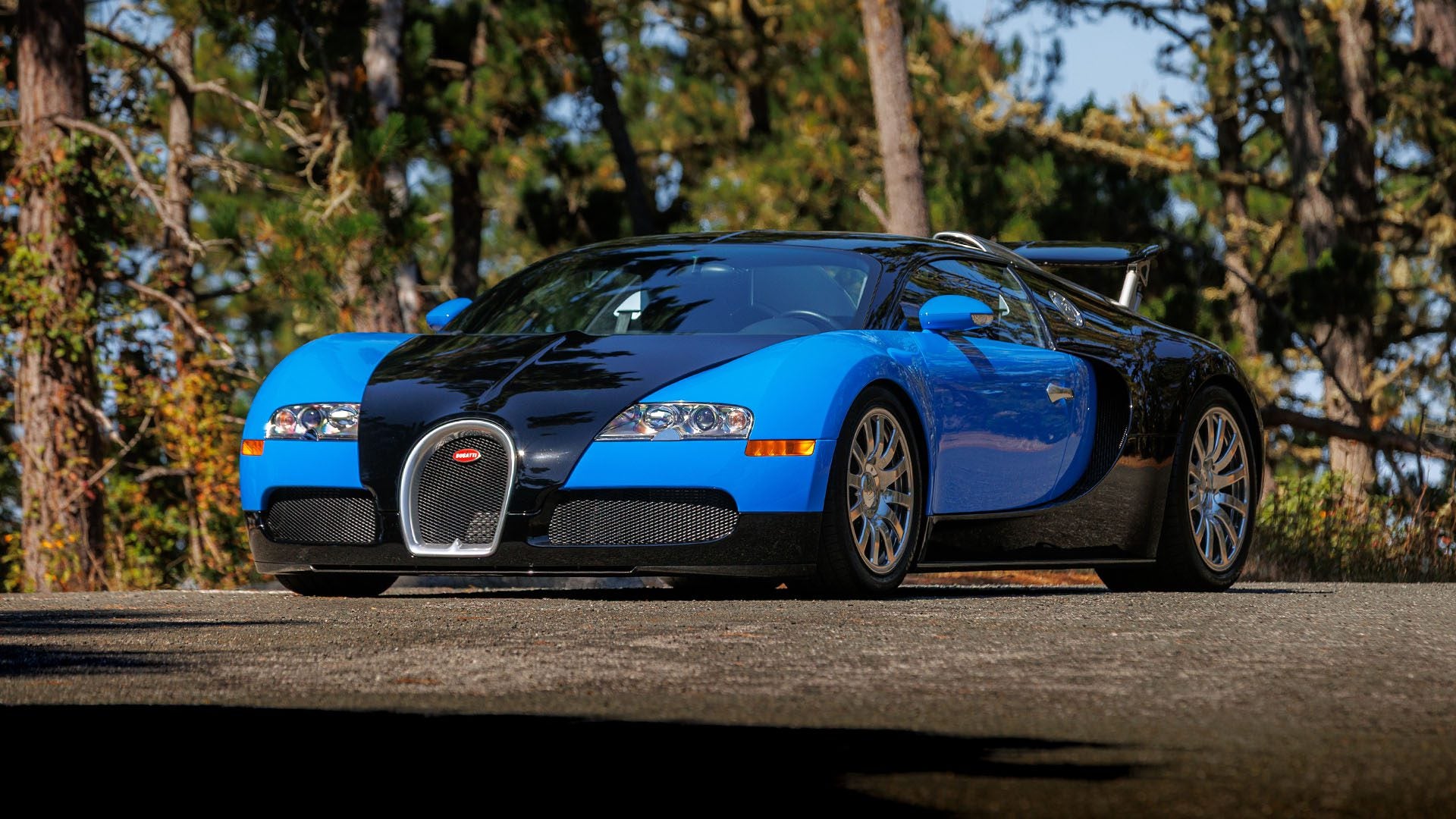 A blue and black Bugatti Veyron sports car is parked on a paved road with trees in the background, ready to turn heads at Broad Arrow’s Las Vegas 2025 event.
