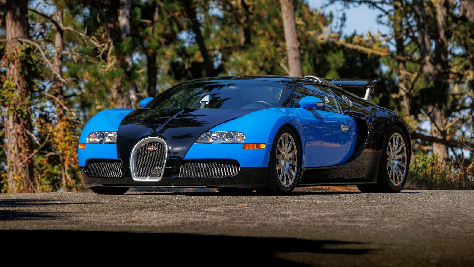 A blue and black Bugatti Veyron sports car is parked on a paved road with trees in the background, ready to turn heads at Broad Arrow’s Las Vegas 2025 event.