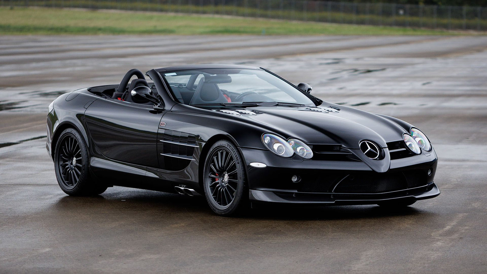 A 2009 Mercedes-Benz SLR McLaren convertible is parked on a wet concrete surface, with a grassy area and fence in the background, possibly awaiting its spotlight at Broad Arrow Las Vegas 2025.