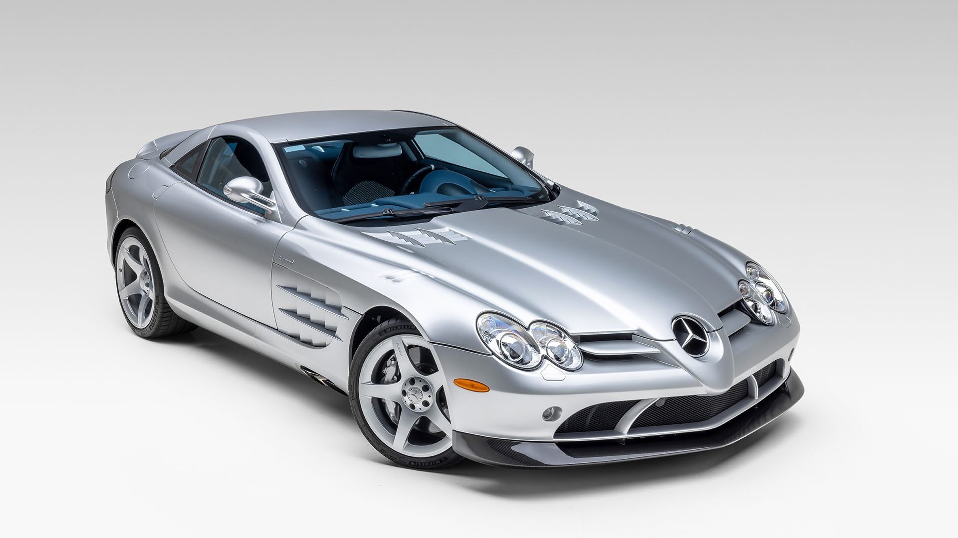A silver Mercedes-Benz SLR McLaren coupe is parked in a studio with a plain white background, reminiscent of the elegance seen at Broad Arrow’s Las Vegas 2025 event.