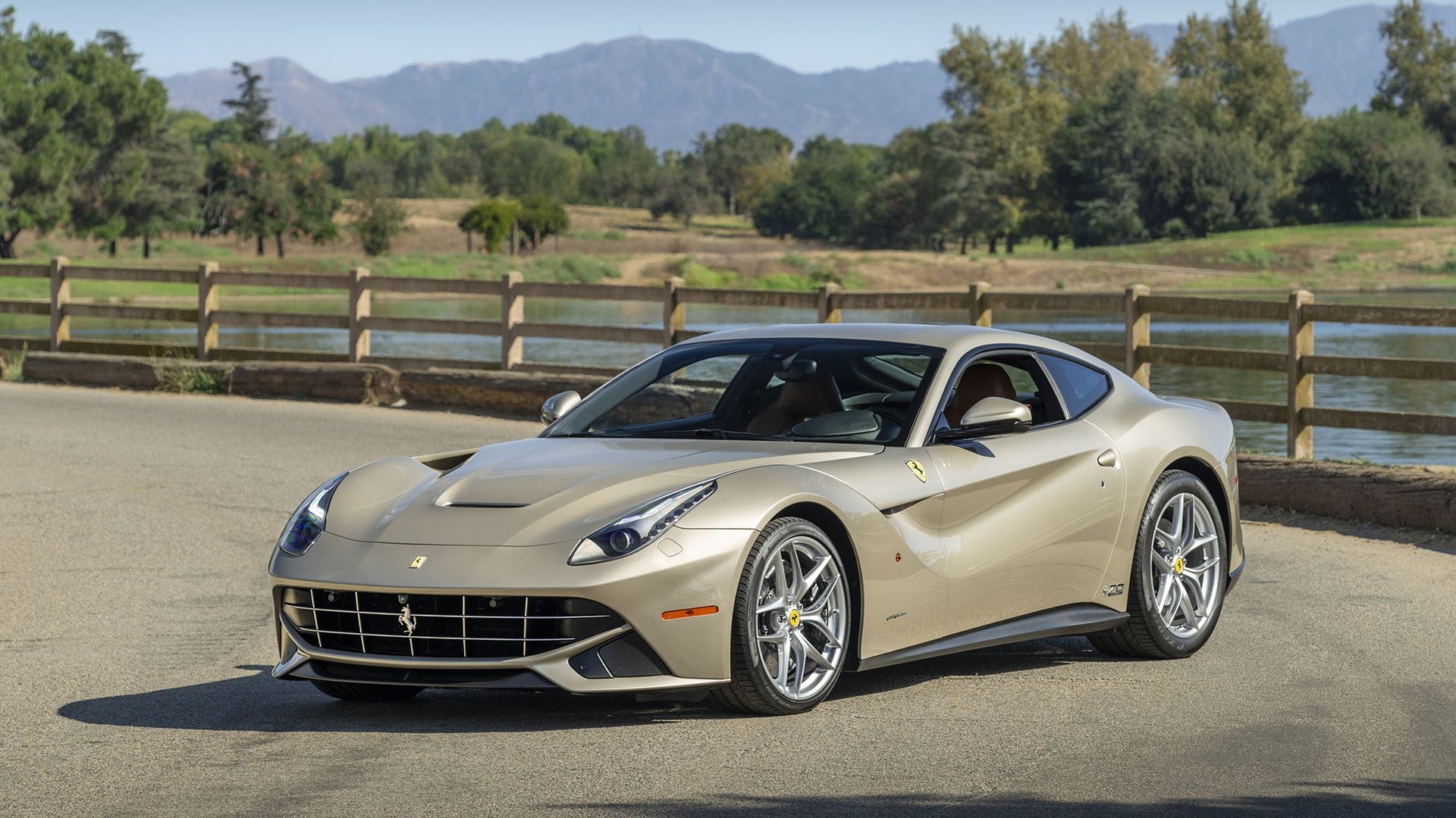 A beige Ferrari F12berlinetta 70th Anniversary Edition is parked on a paved road beside a wooden fence, with trees and mountains in the background, capturing a timeless scene worthy of Las Vegas 2025.