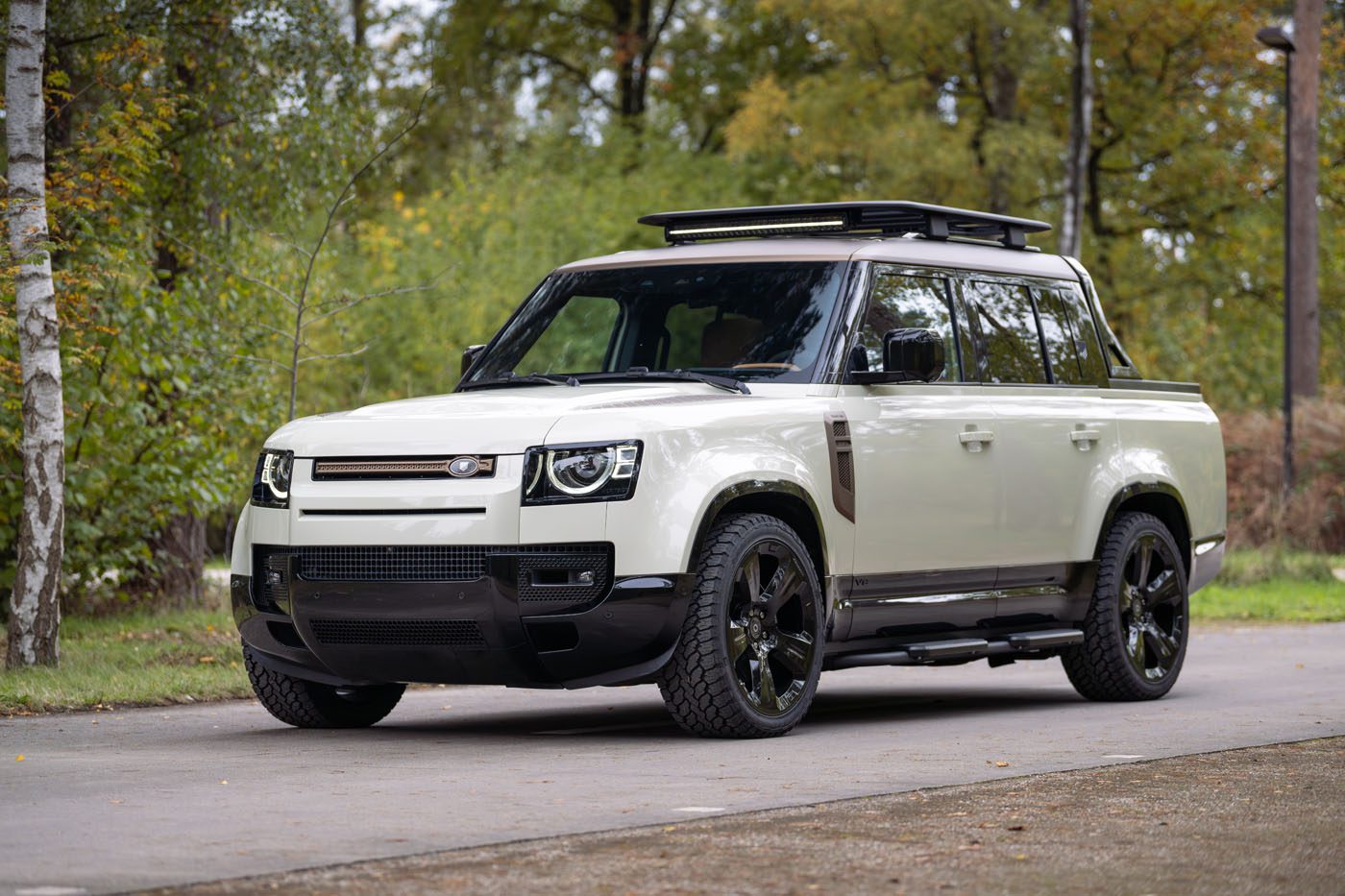 A white Defender Pickup with black wheels and a roof rack from Heritage Customs is parked on a paved road, surrounded by greenery and trees.