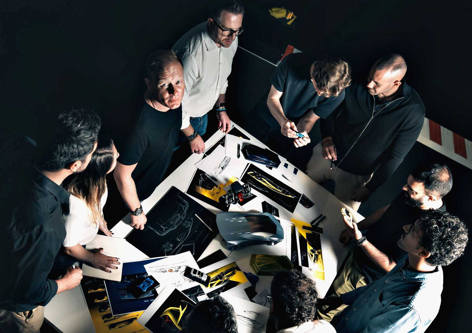 A group of people stands around a table covered with car design sketches, models, and color samples, engaged in discussion—illustrating how Centro Stile is shaping the future of Lamborghini design.