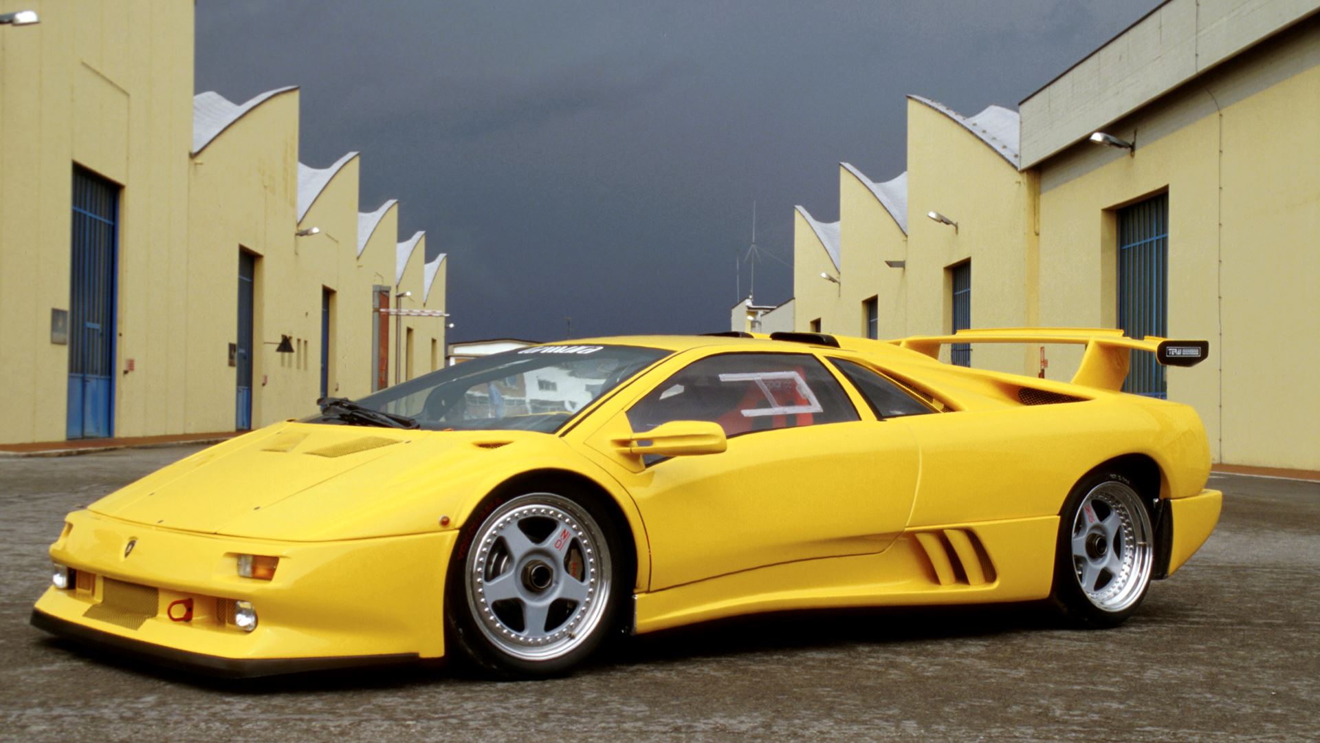 A yellow Lamborghini Diablo—celebrating 35 years as the last old-school analog Raging Bull—stands parked between yellow industrial buildings with blue doors, its legendary roar echoing under a cloudy sky.