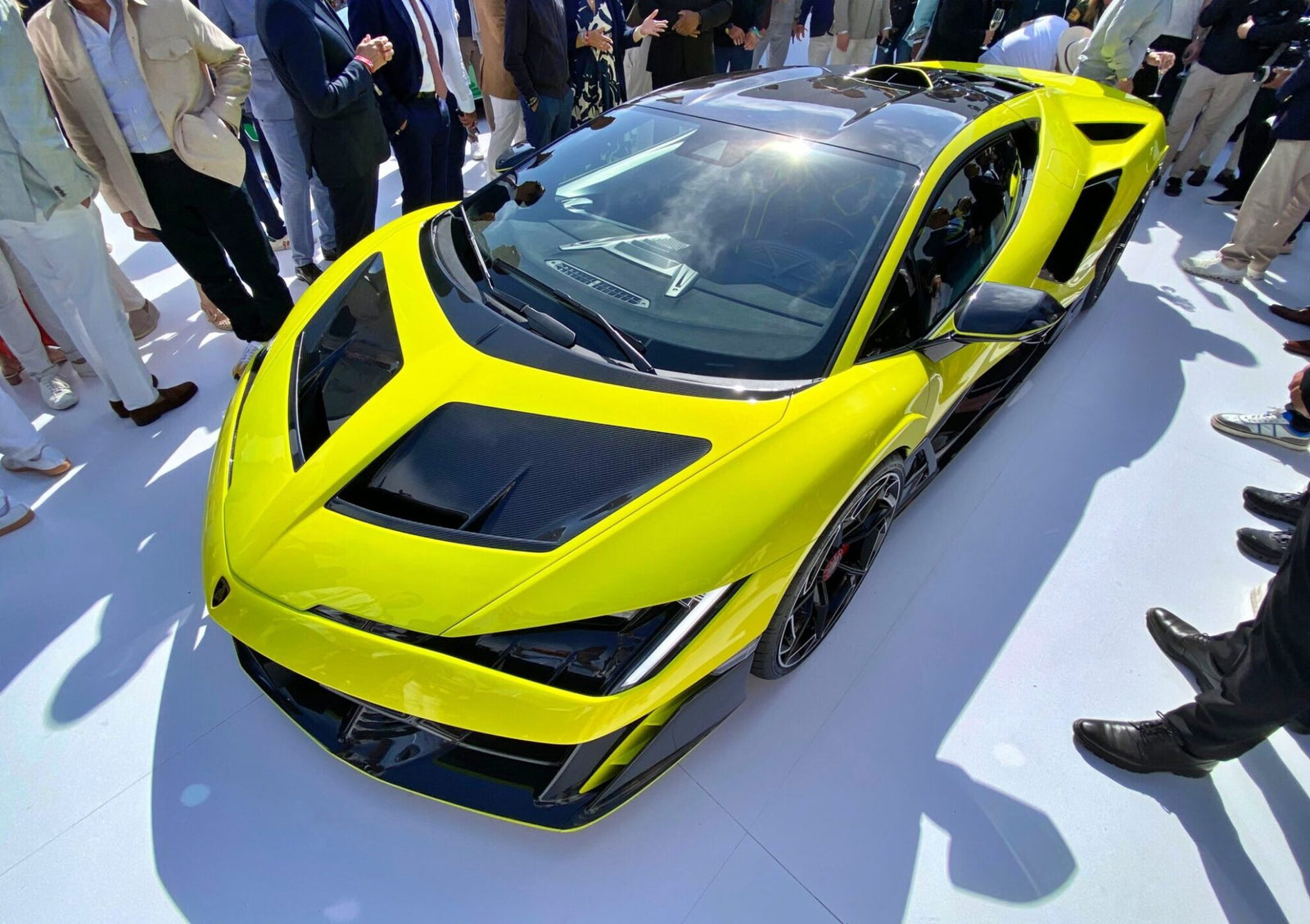 A bright yellow Lamborghini V12 sports car on display, surrounded by a crowd of people, viewed from above—a true collector’s dream.