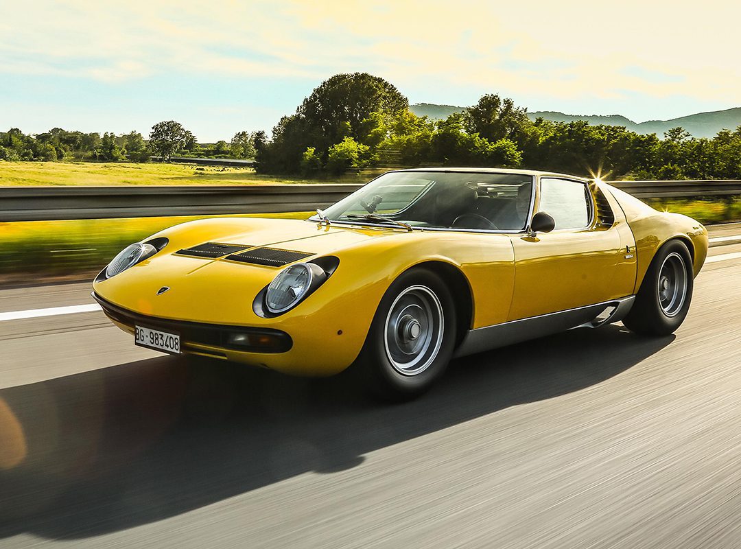 A yellow Lamborghini, celebrated in automotive history and restored by Polo Storico, drives on a highway with trees and mountains in the background on a sunny day.