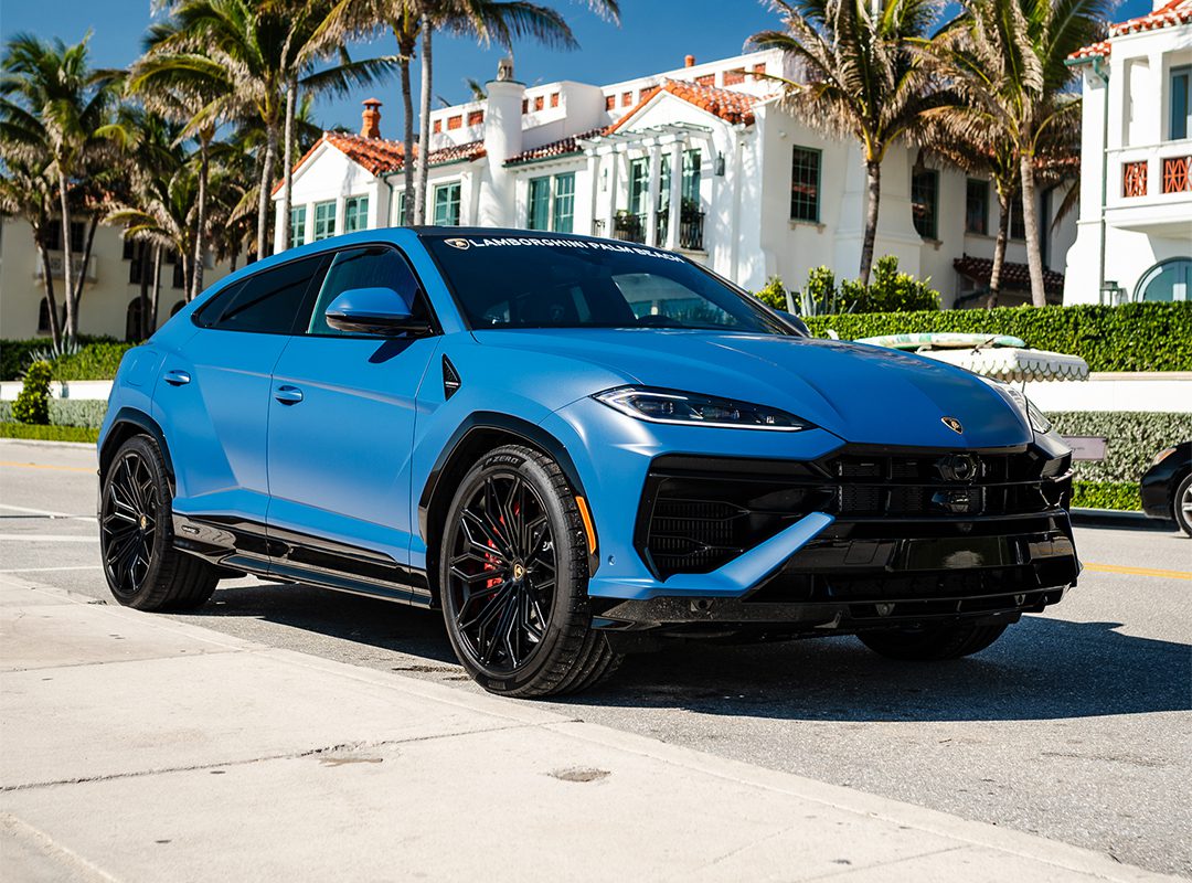 A matte blue Lamborghini Urus SE is parked on a sunny street lined with palm trees and upscale houses, showcasing the luxury of Lamborghini Palm Beach—imagine arriving home in style with a 12-Month Lease.