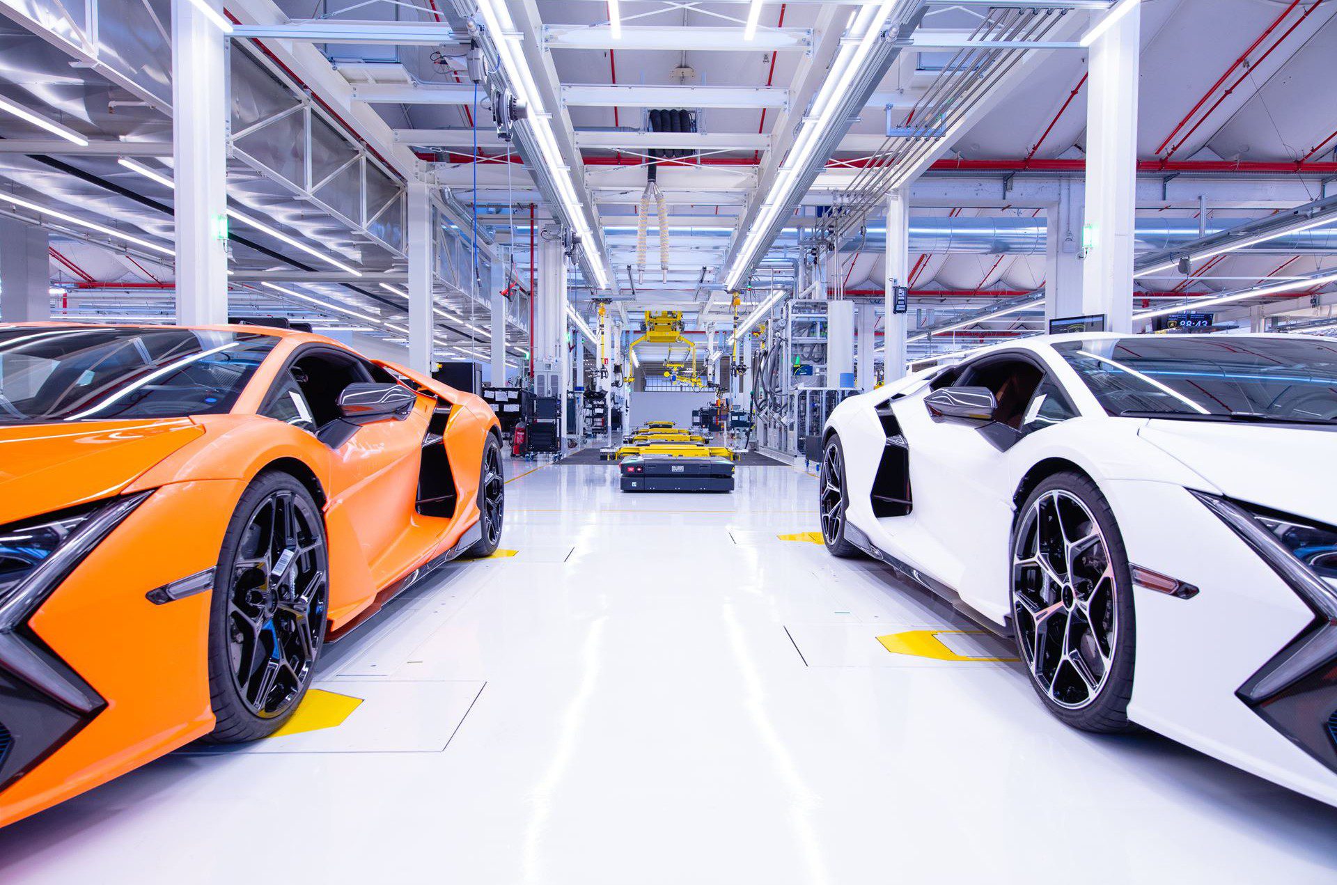 Orange and white sports cars are parked inside a bright, modern automotive manufacturing facility, reflecting the innovation behind Automobili Lamborghini Continues To See Growth In Q1 2025. Machinery and equipment fill the background.