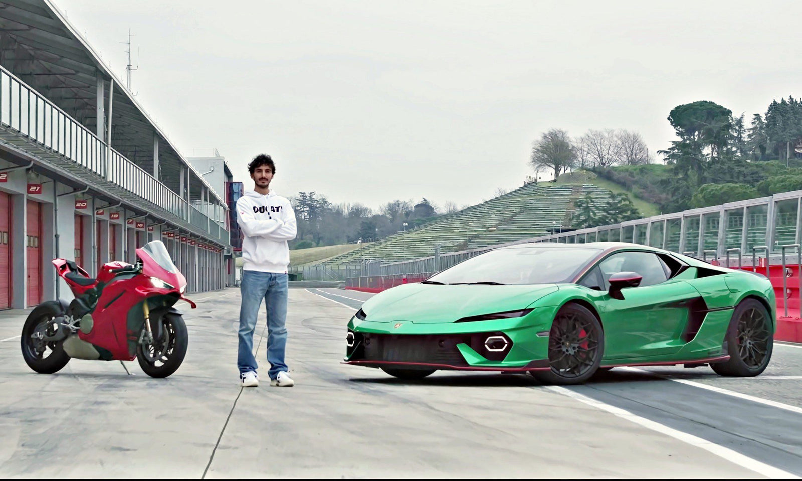 A person stands between a red motorcycle and a green sports car on a racetrack, echoing what two-time MotoGP champ Francesco Bagnaia makes of the Lamborghini Temerario, with empty stands and overcast skies in the background.