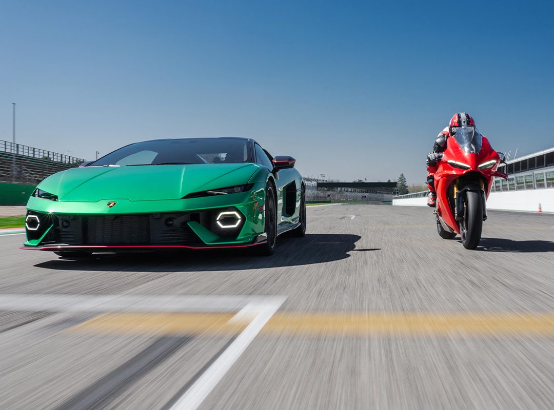 A green Lamborghini Temerario and a red Ducati Panigale V4 drive head-to-head on a racetrack under a clear blue sky.