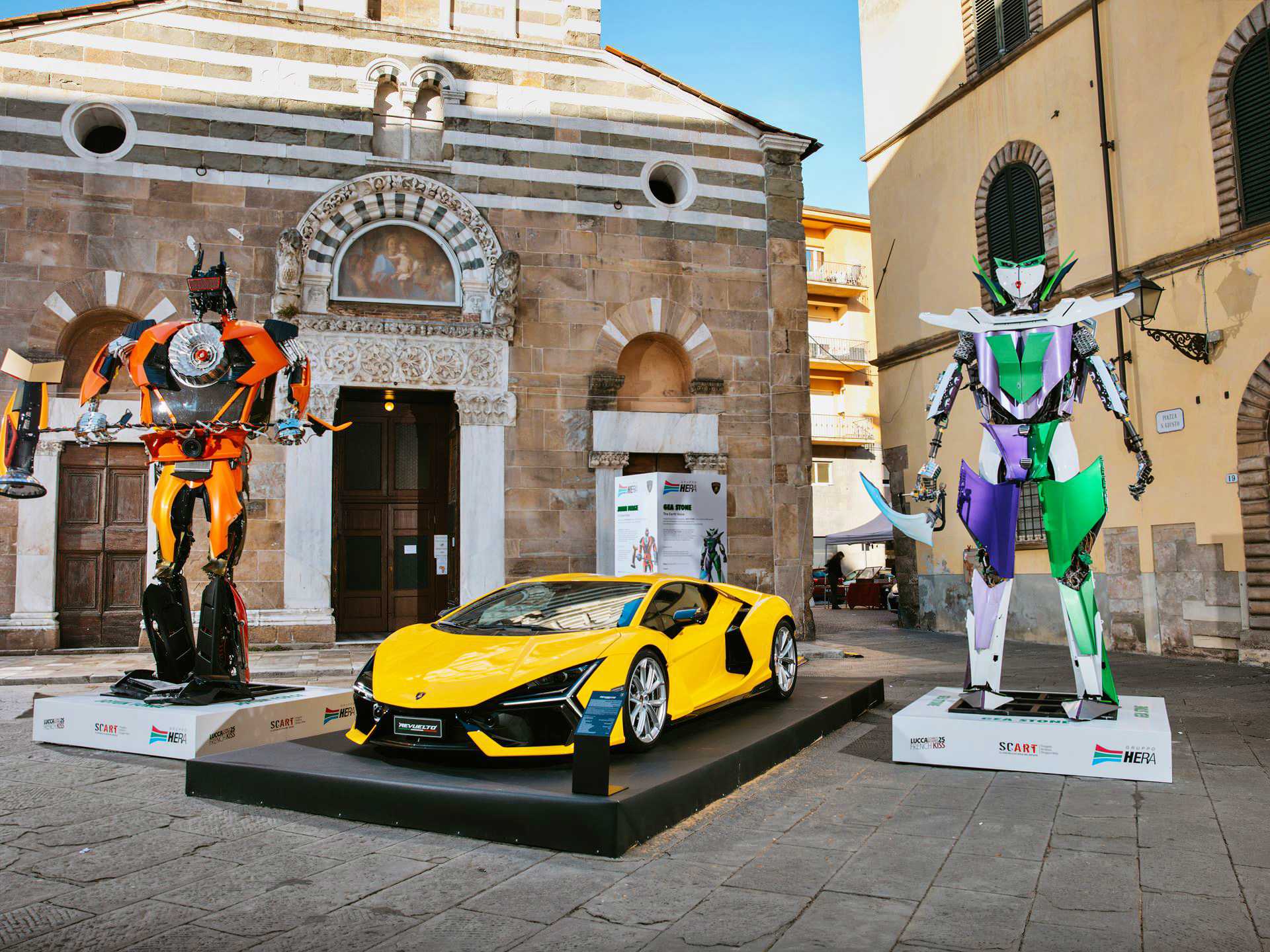 A yellow sports car is displayed between two giant robot sculptures at Lucca Comics & Games, where Lamborghini turns factory waste into impressive robots, set against the backdrop of historic buildings in an outdoor plaza.