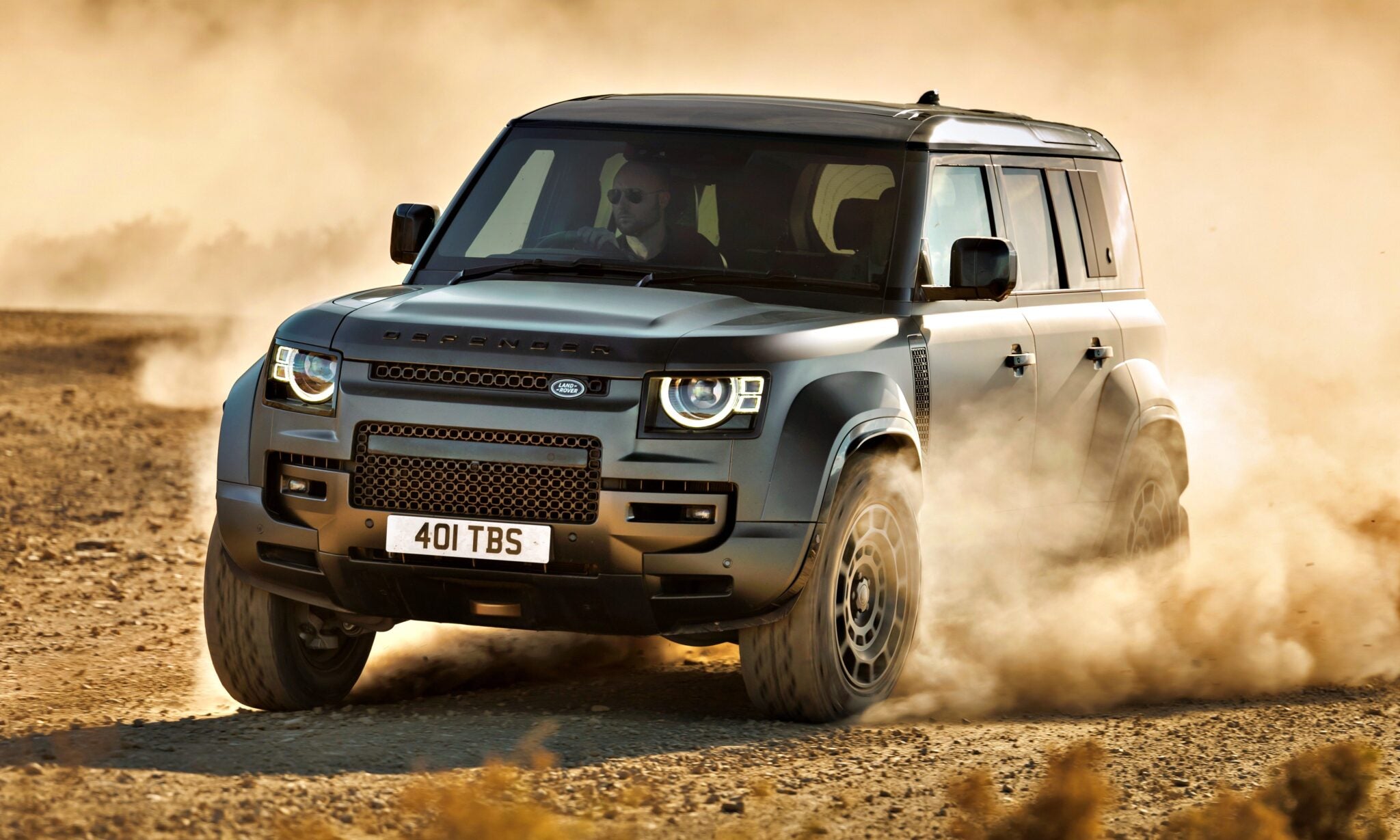 A dark gray Land Rover Defender, a symbol of Here's What's Driving JLR's Best Annual Profits In Nearly A Decade, speeds along a dusty off-road trail, kicking up dirt clouds with its driver clearly visible inside.
