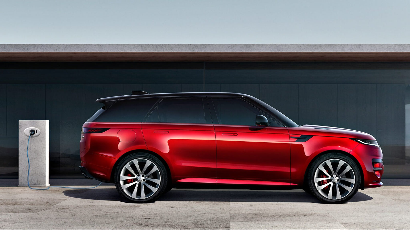 A red SUV, the 2025 Land Rover Range Rover Sport Dynamic SE PHEV, is parked next to an electric vehicle charging station on a concrete surface, with a modern building in the background.