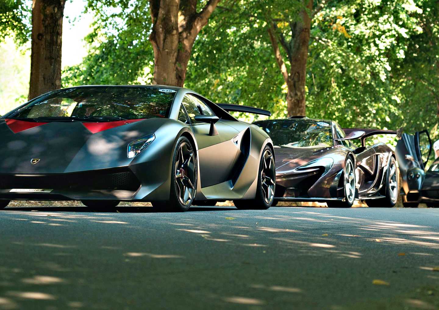Two high-performance sports cars, including a street-legal Lamborghini Sesto Elemento in matte black, are parked under leafy trees on a sunlit road.
