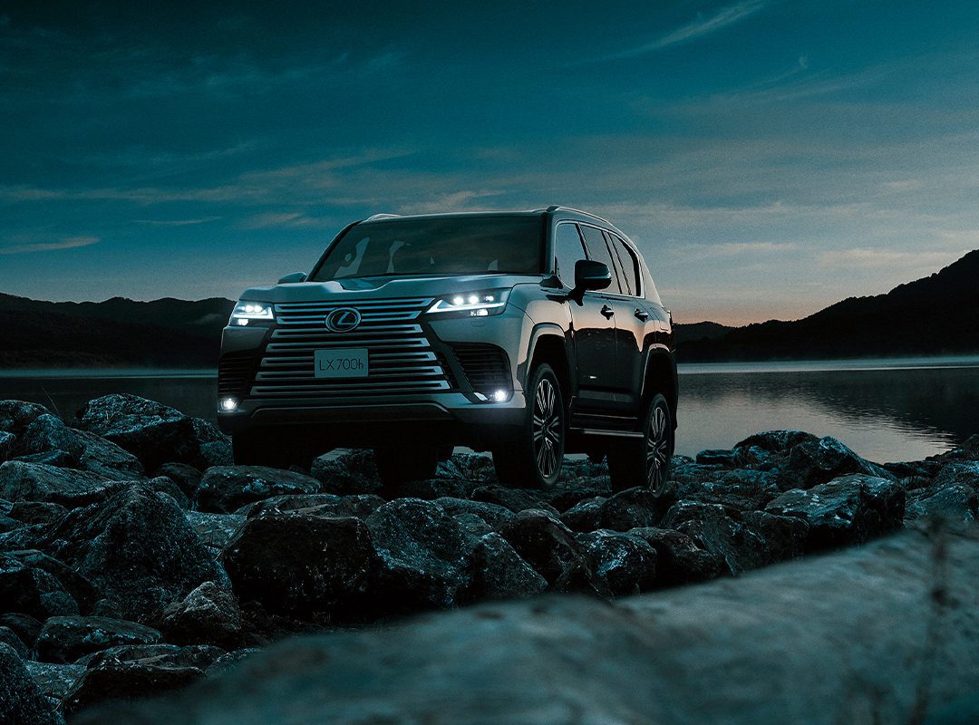 A Lexus SUV, embodying Expedition Elegance, sits with headlights aglow on rocky terrain by a lake at dusk, framed by hills and a darkening sky.