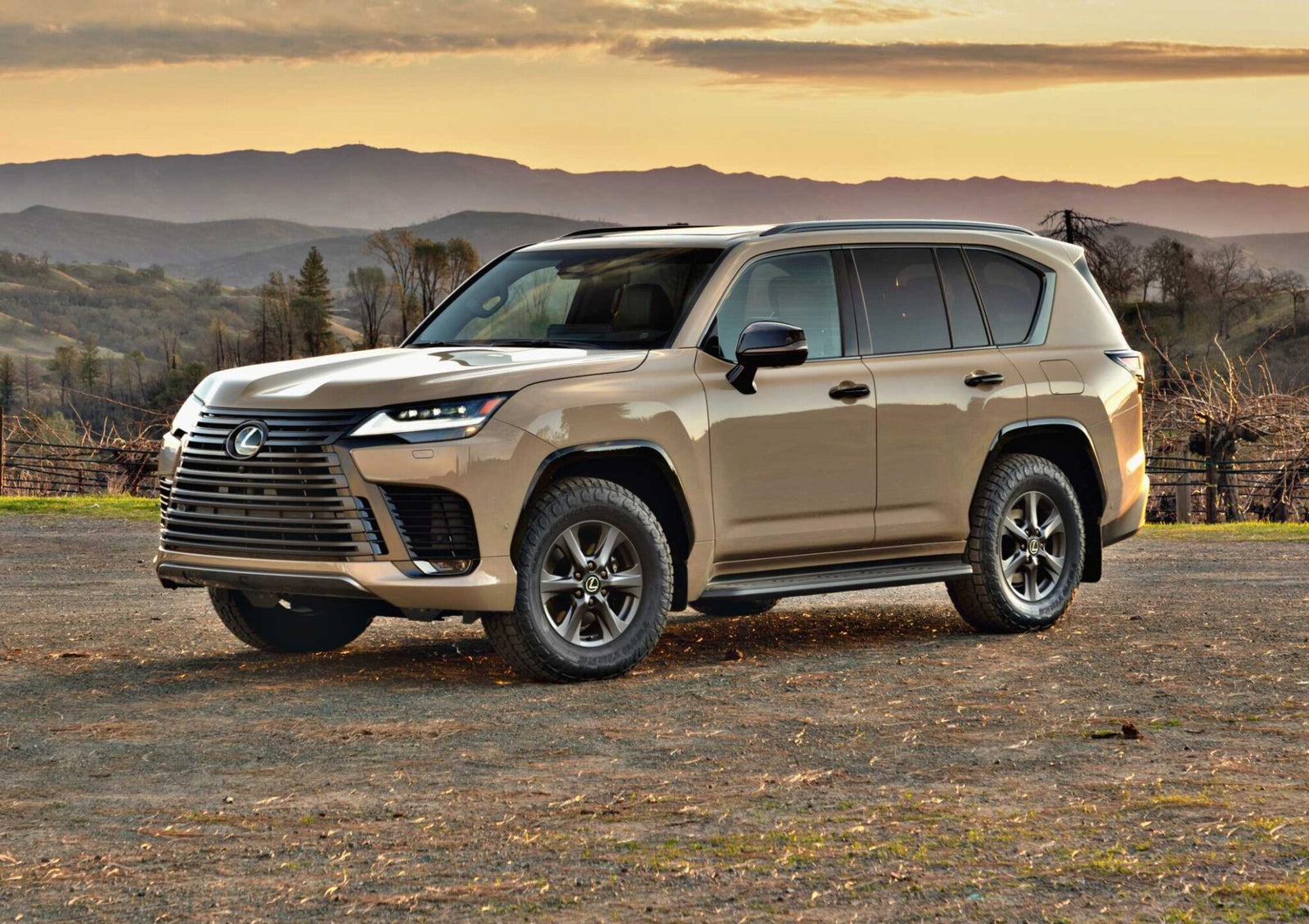 A beige Lexus SUV, the 2025 Lexus LX 700h Overtrail 4WD, is parked on dirt ground with mountains and trees in the background at sunset—showcasing its highly capable yet moderately practical design.