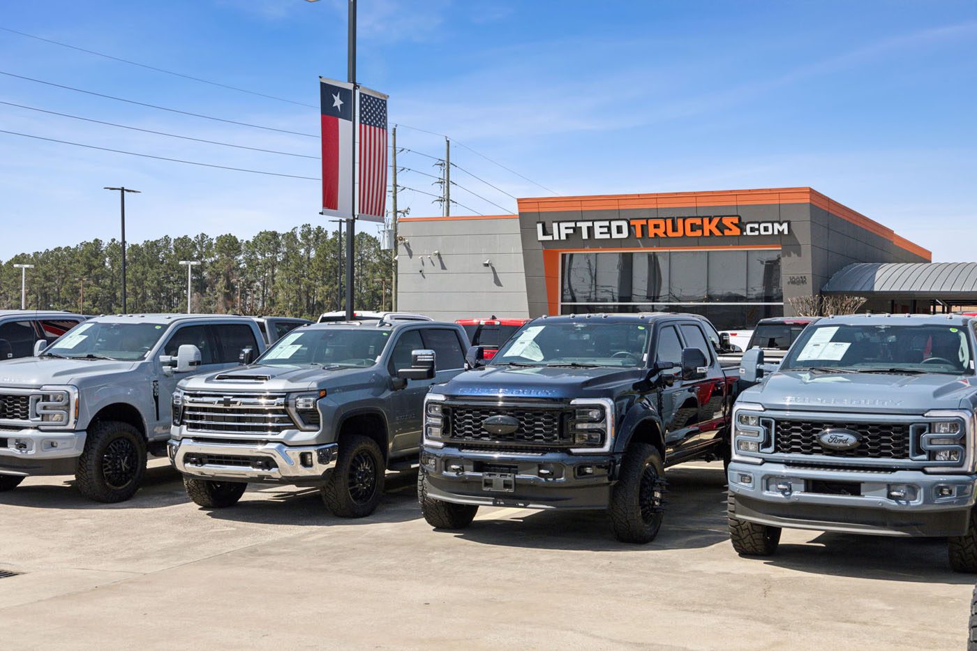 Four large pickup trucks are parked in front of a dealership with a "LIFTEDTRUCKS.com" sign, proudly showcasing America’s Original Lifted Truck Dealer, with a Texas flag visible in the background.