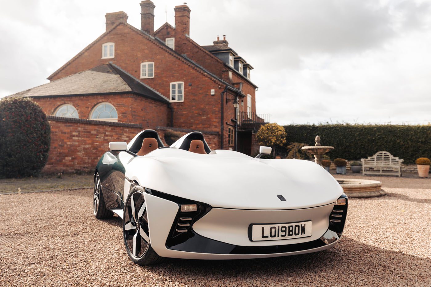 A white, two-seater sports car with license plate LO1980M is parked on a gravel driveway in front of a large brick house.