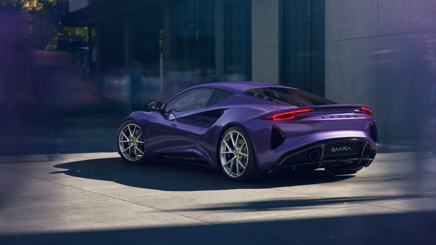 A purple Lotus Emira sports car is parked on a city street beside a modern building, with blurred figures in the background.