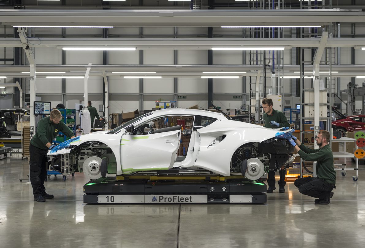 Three workers assemble a white Lotus sports car on a factory floor, with the car body elevated and wheels not yet attached—a glimpse into U.S. production at the crossroads of innovation and precision craftsmanship.