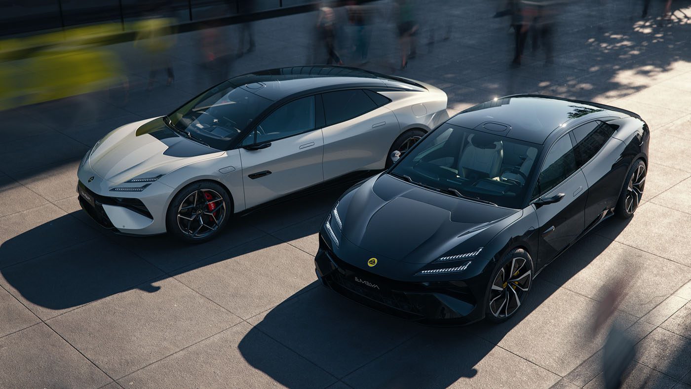 Two sleek sports cars, a white Lotus Emeya and a black Eletre, are parked next to each other on a paved surface, with the blurred motion of people bustling around them.