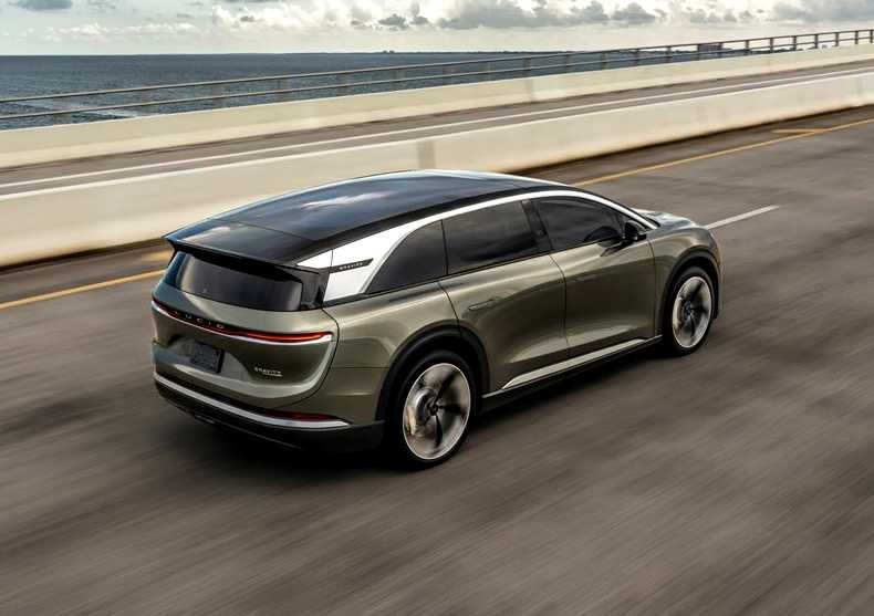 A green electric SUV with impressive autonomy drives on a highway next to the ocean under a partly cloudy sky.
