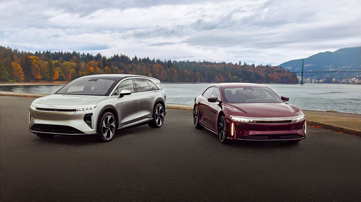 Two modern electric cars, one silver and one maroon, parked side by side on a paved area with a scenic lake, bridge, and autumn trees—reminiscent of Lucid's record Q3 deliveries amid the EV market slowdown.