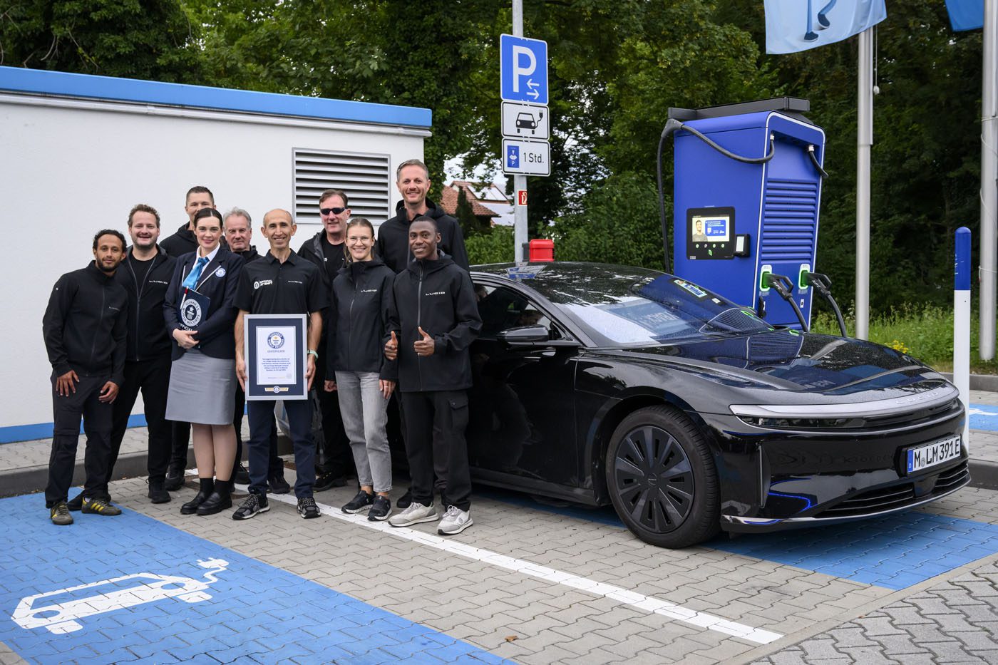 A group of people stand and pose by a black Lucid electric car with a 749-mile EV range at a charging station, holding a certificate—possibly for a Guinness World Record—in a parking lot with electric vehicle markings.