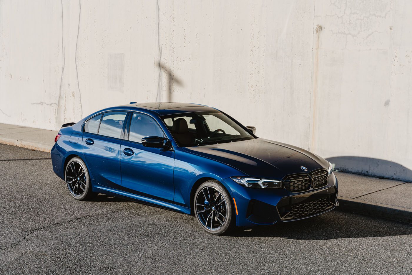 A blue BMW M340i xDrive sedan, 50 Jahre Edition, is parked on a sunlit street next to a light-colored concrete wall.