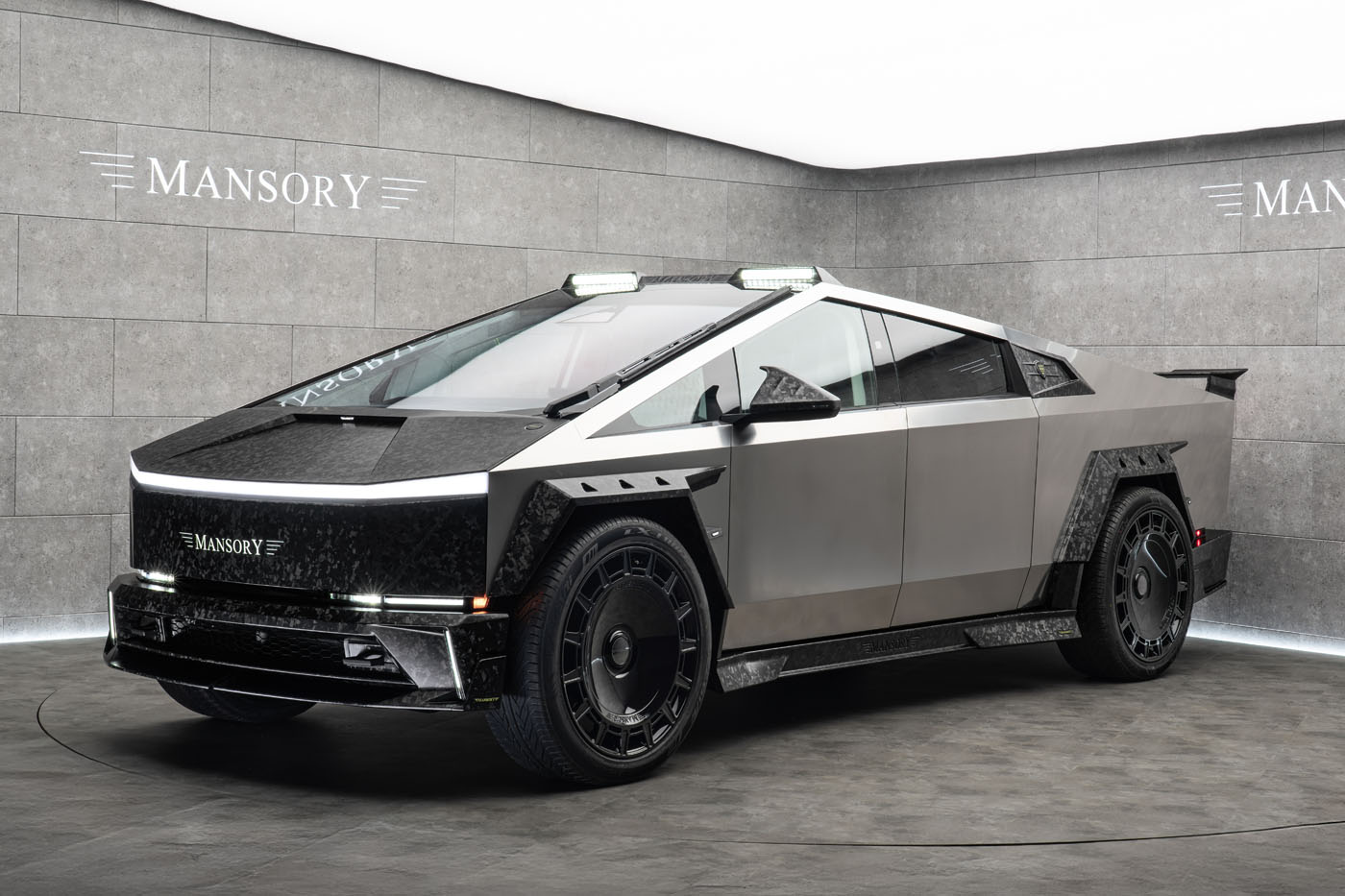 The Custom Tesla Cybertruck, with its futuristic silver and black angular design and subtle elongation, is prominently showcased in a showroom adorned with "Mansory" branding on the walls.