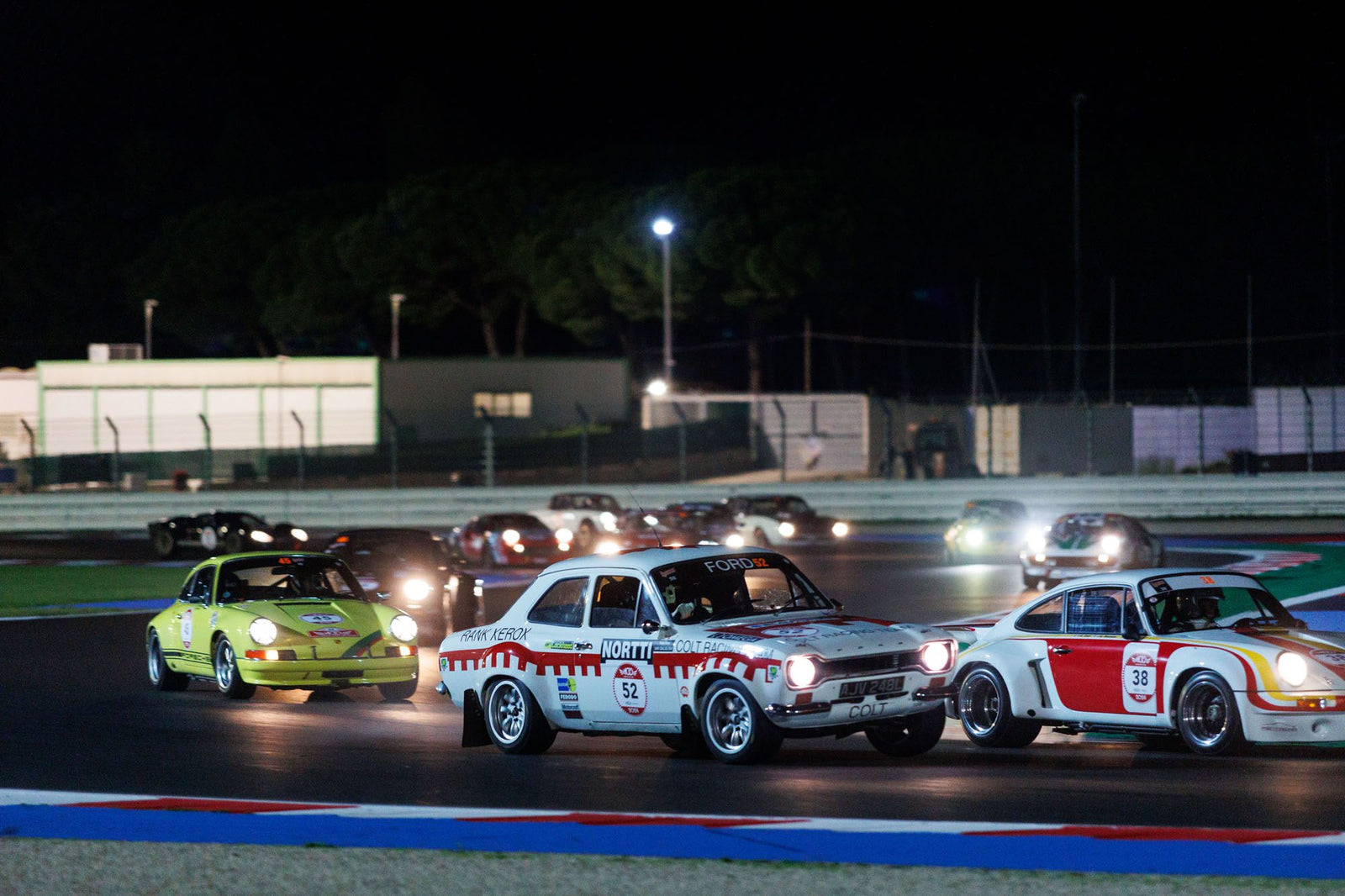 A group of vintage race cars competes on a dimly lit track during the Modena Cento Ore night race, with blurred trees and buildings in the background.