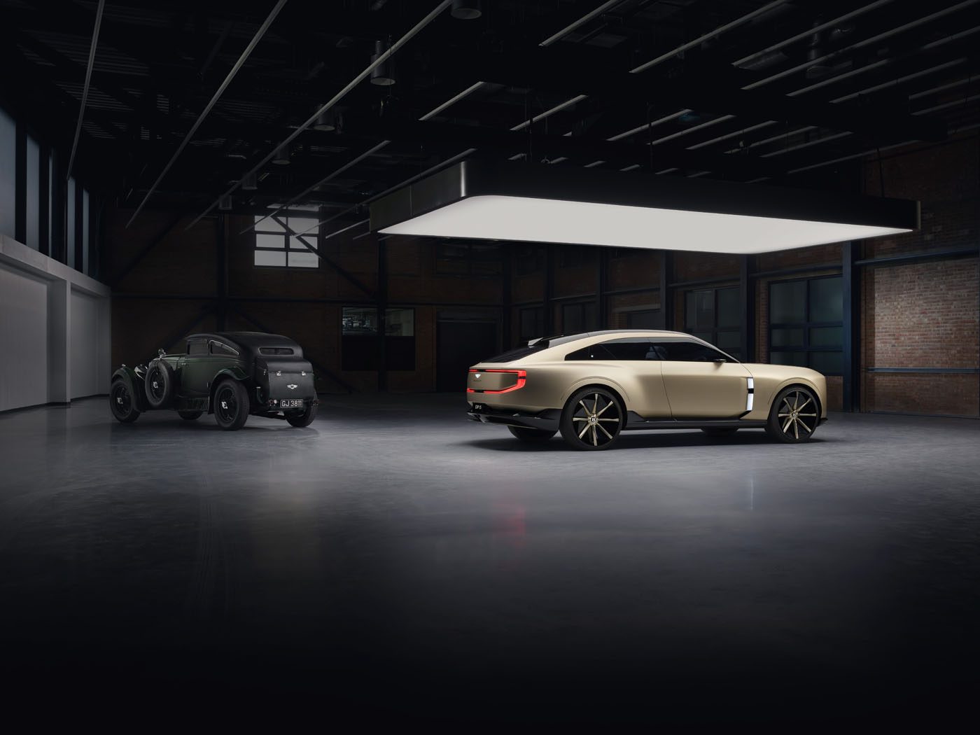 A vintage black car and a modern beige Bentley EXP 15 Concept are parked side by side in a spacious, dimly lit industrial garage, capturing the spirit of Monterey Car Week.