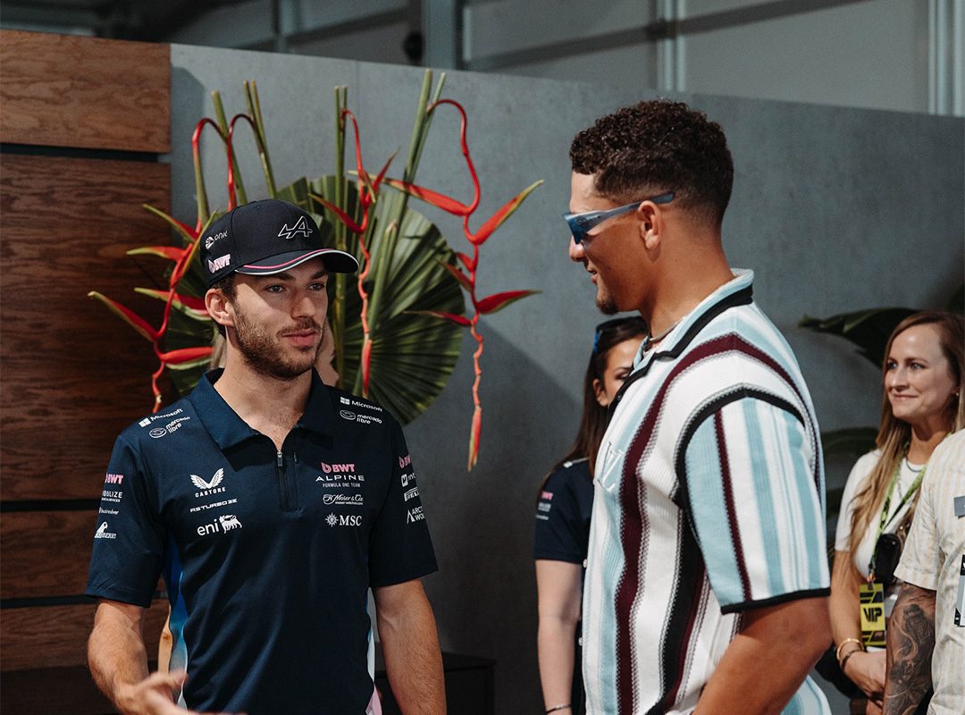 Two men stand and talk indoors at the Miami Grand Prix—one in a racing team shirt and cap, the other in sunglasses and a striped shirt. Three people are visible in the background during this F1 2024 Celebrity Sightings moment.