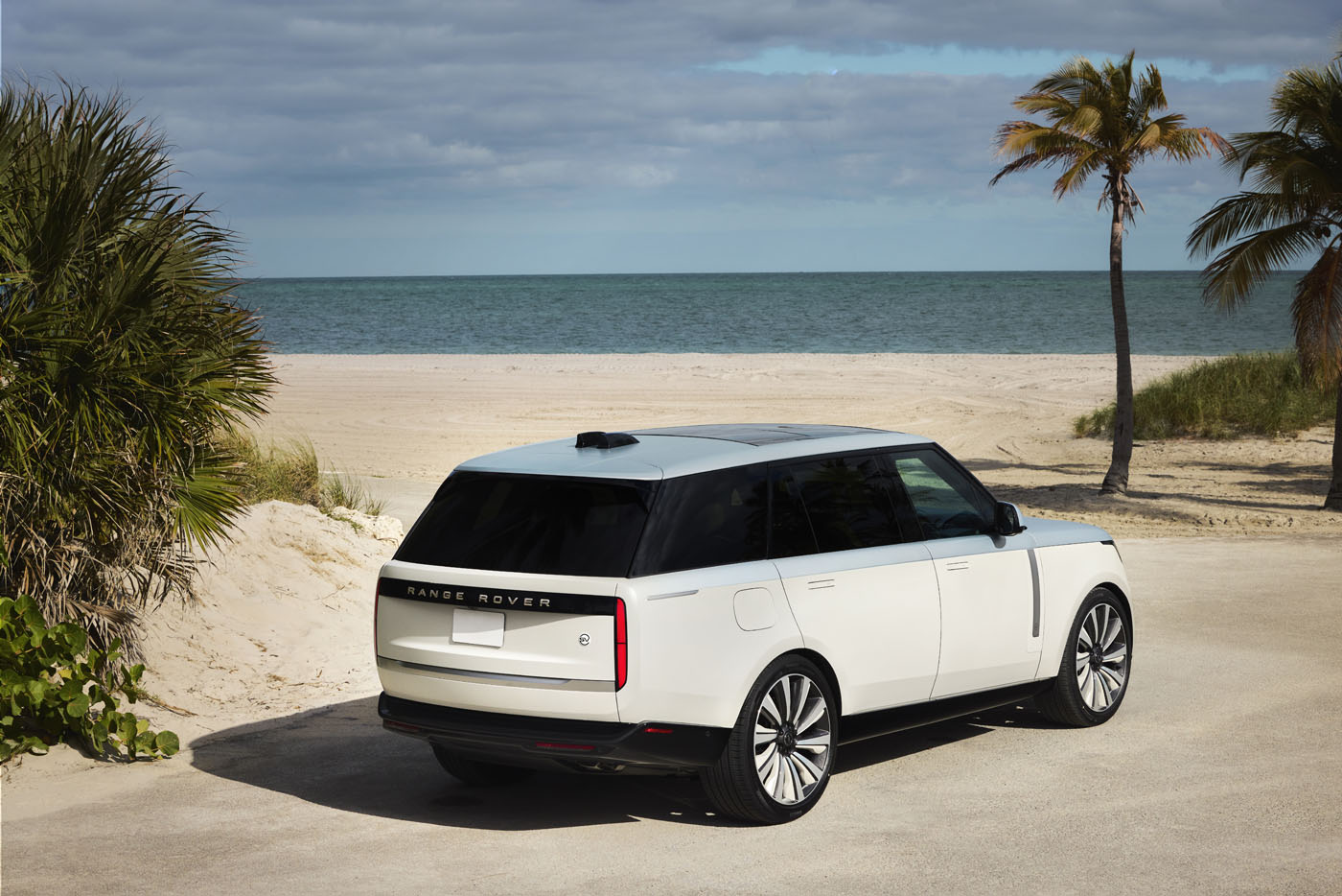 The white Range Rover SV Candeo is elegantly parked on a sandy beach with palm trees, overlooking a calm ocean under a cloudy sky.