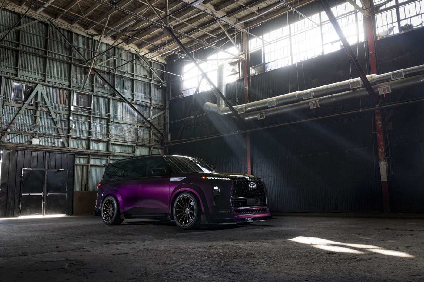 A dark purple QX80 R-Spec SUV is parked inside an old, industrial warehouse with sunlight streaming through large windows above, hinting at its impressive 1,000HP GT-R NISMO engine under the hood.