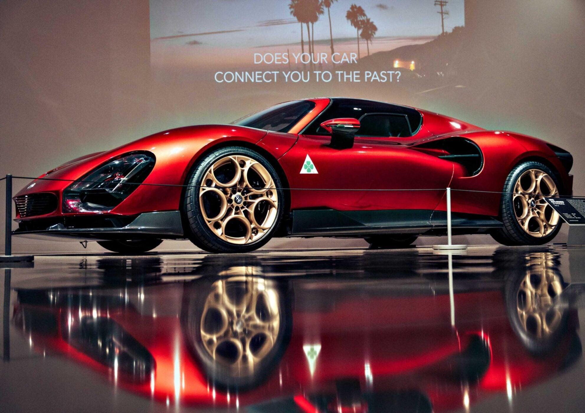 A red sports car with gold rims is displayed indoors at the Petersen Automotive Museum. A sign in the background reads, "DOES YOUR CAR CONNECT YOU TO THE PAST?.