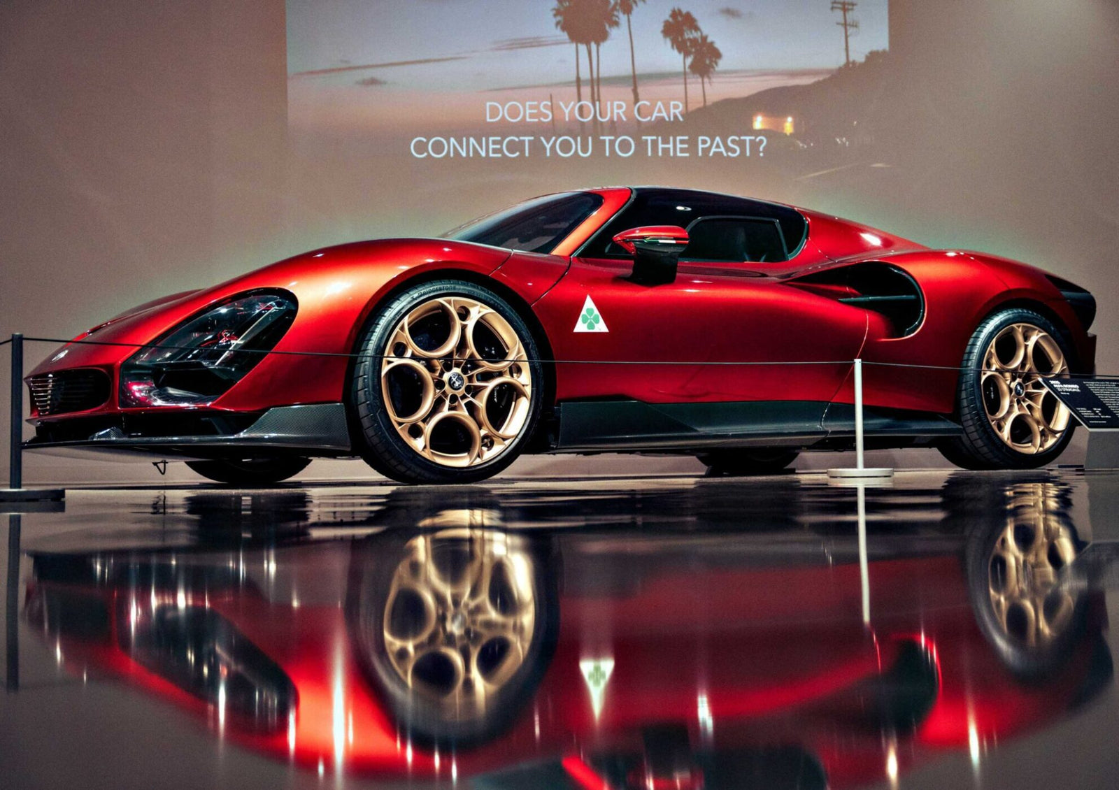 A red sports car with gold rims is displayed indoors at the Petersen Automotive Museum. A sign in the background reads, "DOES YOUR CAR CONNECT YOU TO THE PAST?.