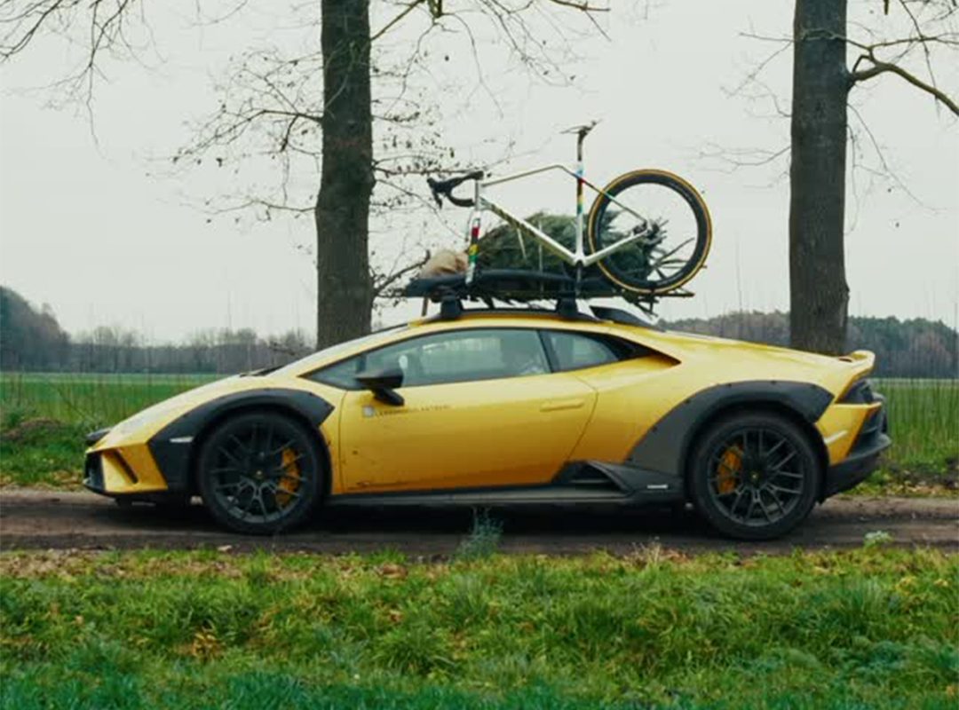 An Off-Road Lamborghini, with a bicycle and a small Christmas tree secured to its roof, is parked on a rural dirt road next to trees and grass—ready for a World Champion Cyclist’s adventurous holiday getaway.