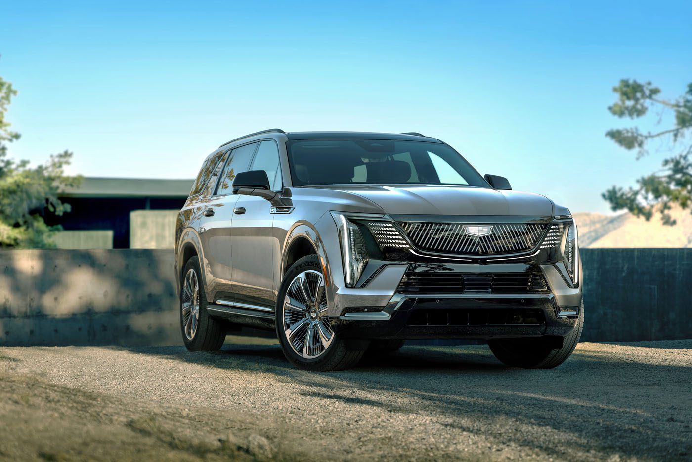 A sleek silver 2026 Escalade IQL sits on a gravel surface under a clear blue sky, with trees and a building in the background.