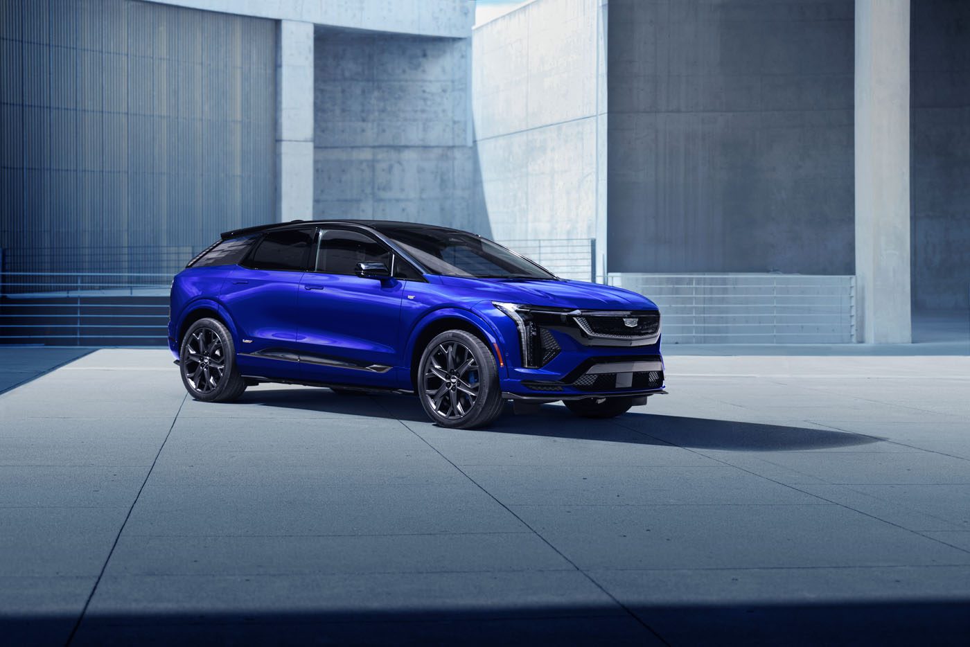 A blue Cadillac Lyriq electric SUV is parked on a concrete surface in front of a modern building with grey walls.