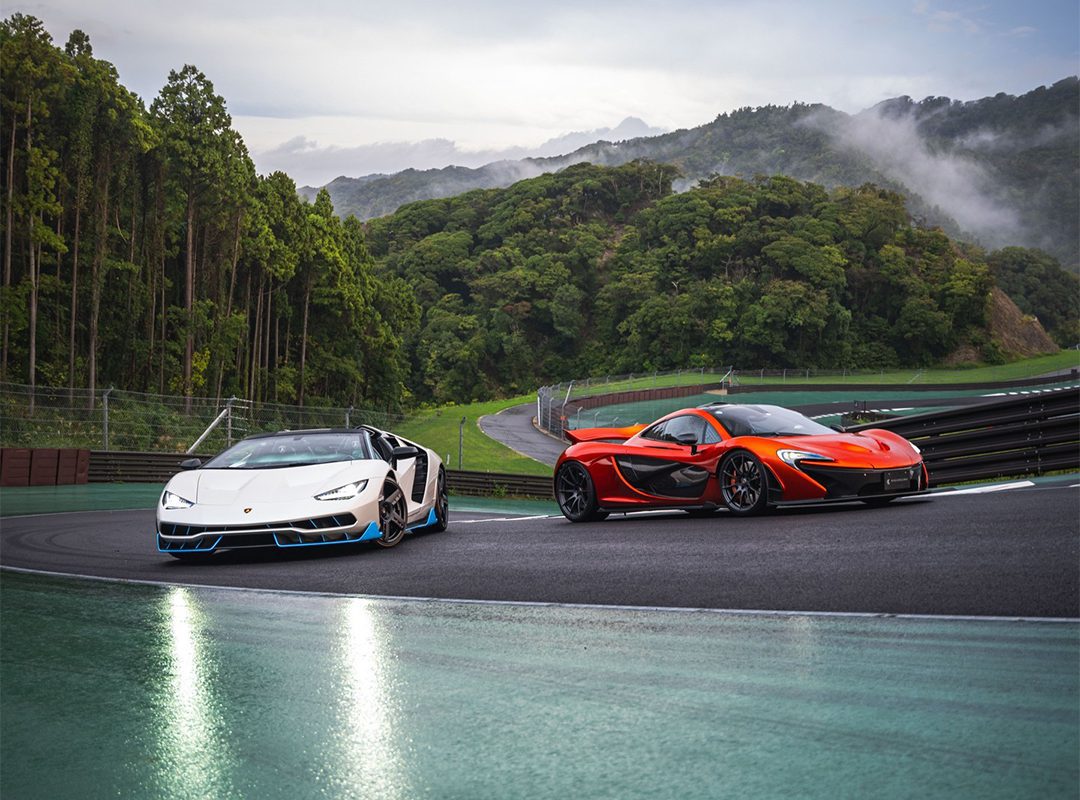 Two sports cars, one white and one red, zip around Asia's first-ever private members-only race circuit at Magarigawa Club. The curved track cuts through lush green hills and trees under a cloudy sky.