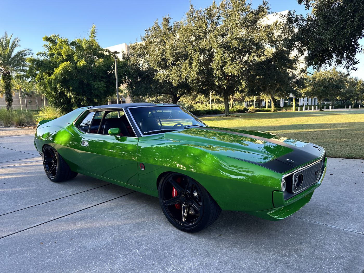 A bright green 1972 AMC Javelin muscle car is parked on a concrete driveway with trees and lawn in the background.