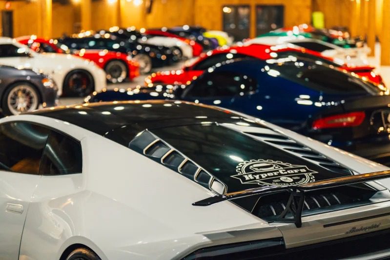 A row of exotic luxury sports cars, including a white car in the foreground with a HyperCars decal, is displayed indoors under warm lighting, offering an unparalleled buying experience.