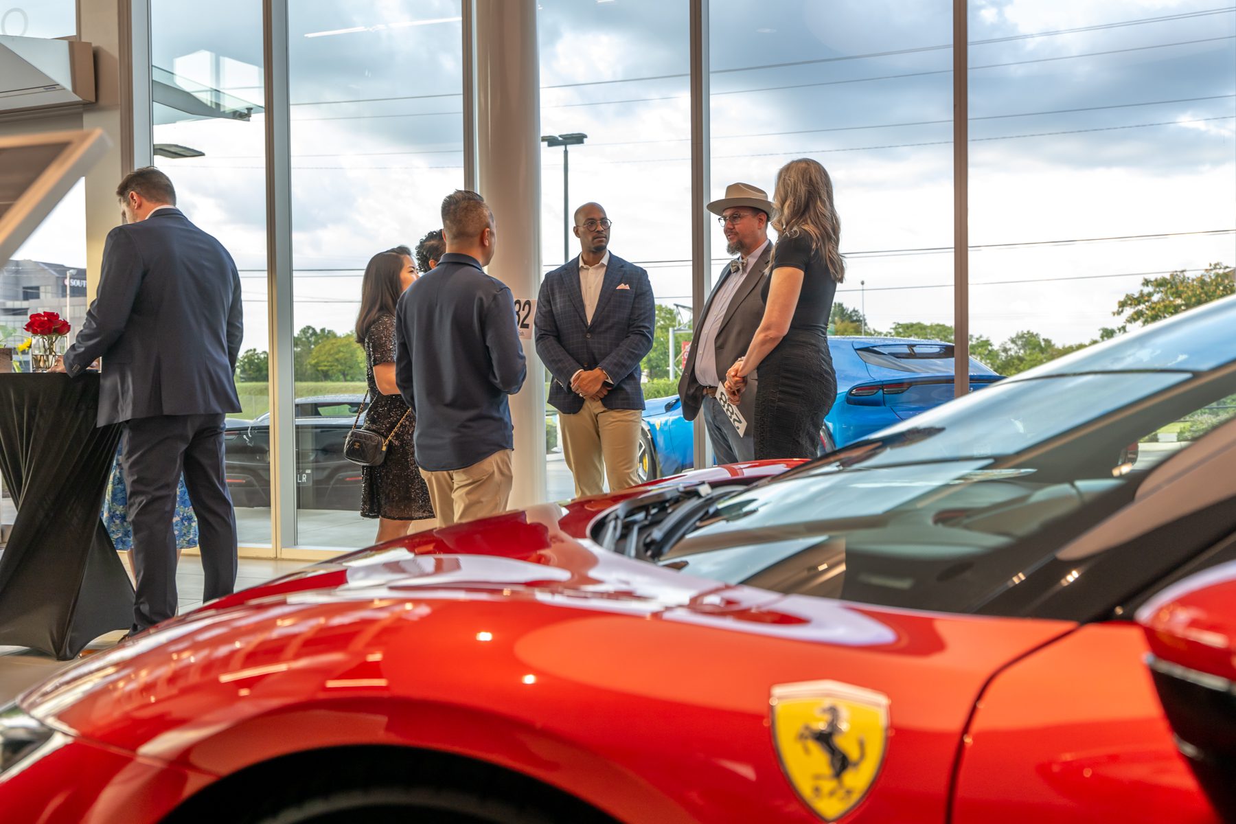 A group of people stand and converse in a car showroom next to a red Ferrari, their wishes in motion as evening light streams through large windows showing cars and trees outside.