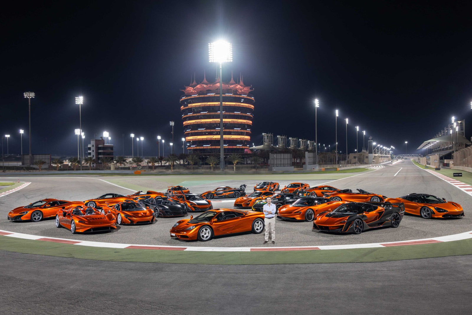 A group of orange sports cars, part of The Most Extraordinary McLaren Collection On The Planet Just Sold To A Single Buyer, is parked on a racetrack at night with an illuminated tower in the background. A person stands in front of the cars.