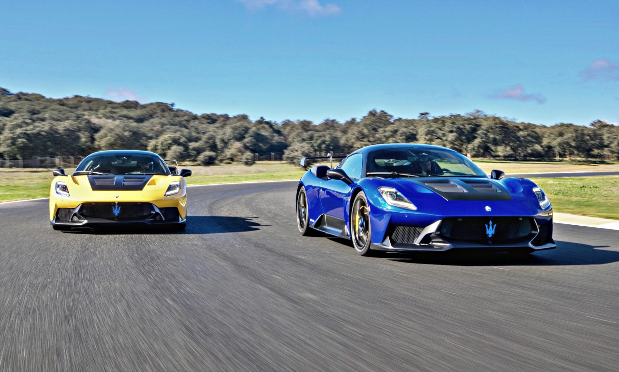 Two Maserati MC20 sports cars, one yellow and one blue, race side by side on a track with trees and blue sky in the background—hinting at how Maserati will take the spotlight at 2025 FuoriConcorso with GT2 Stradale and MCXtrema.