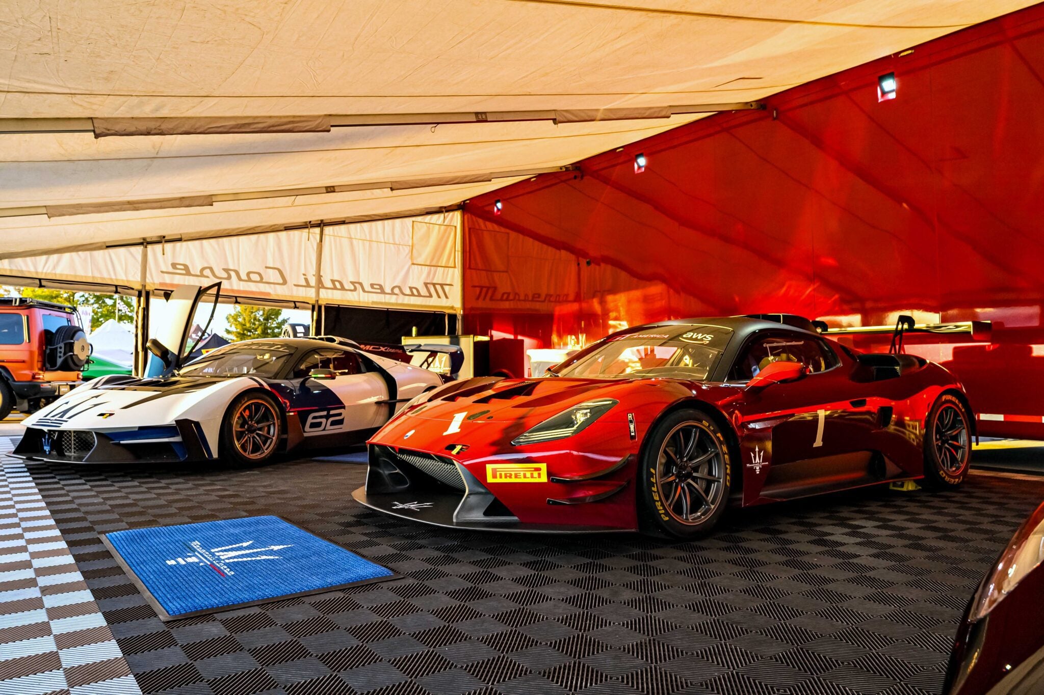 Two Maserati sports cars, one red and one white, are parked inside a tented area with checkered flooring and branded mats as the Maserati GT2 debuts stateside at GT America in Indianapolis.