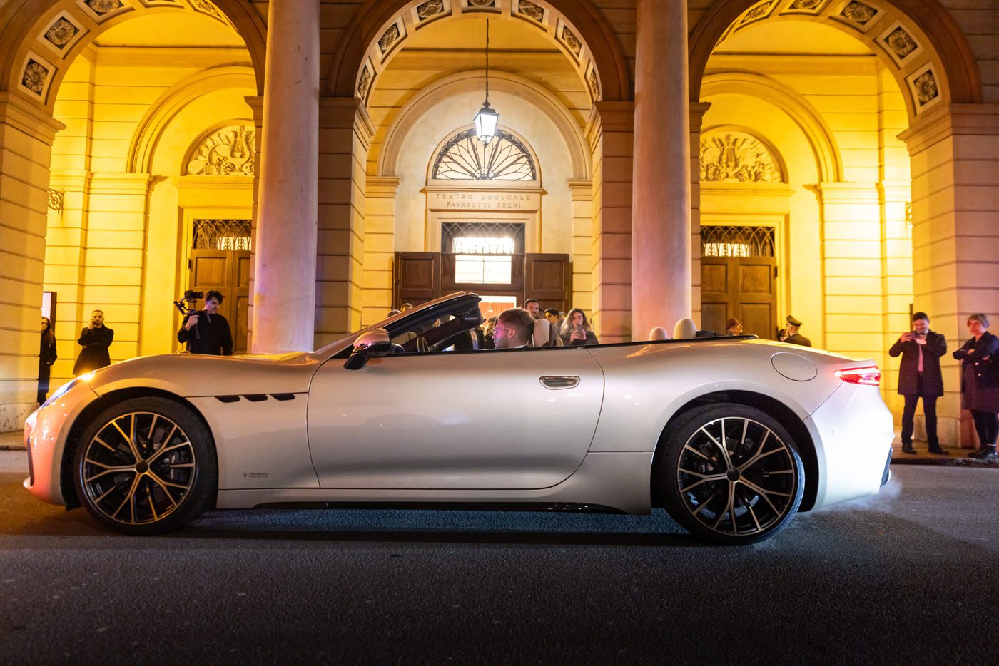 A white Maserati convertible sports car with its top down is parked in front of an illuminated building at night, as several people stand nearby taking photos and admiring its Meccanica Lirica elegance.