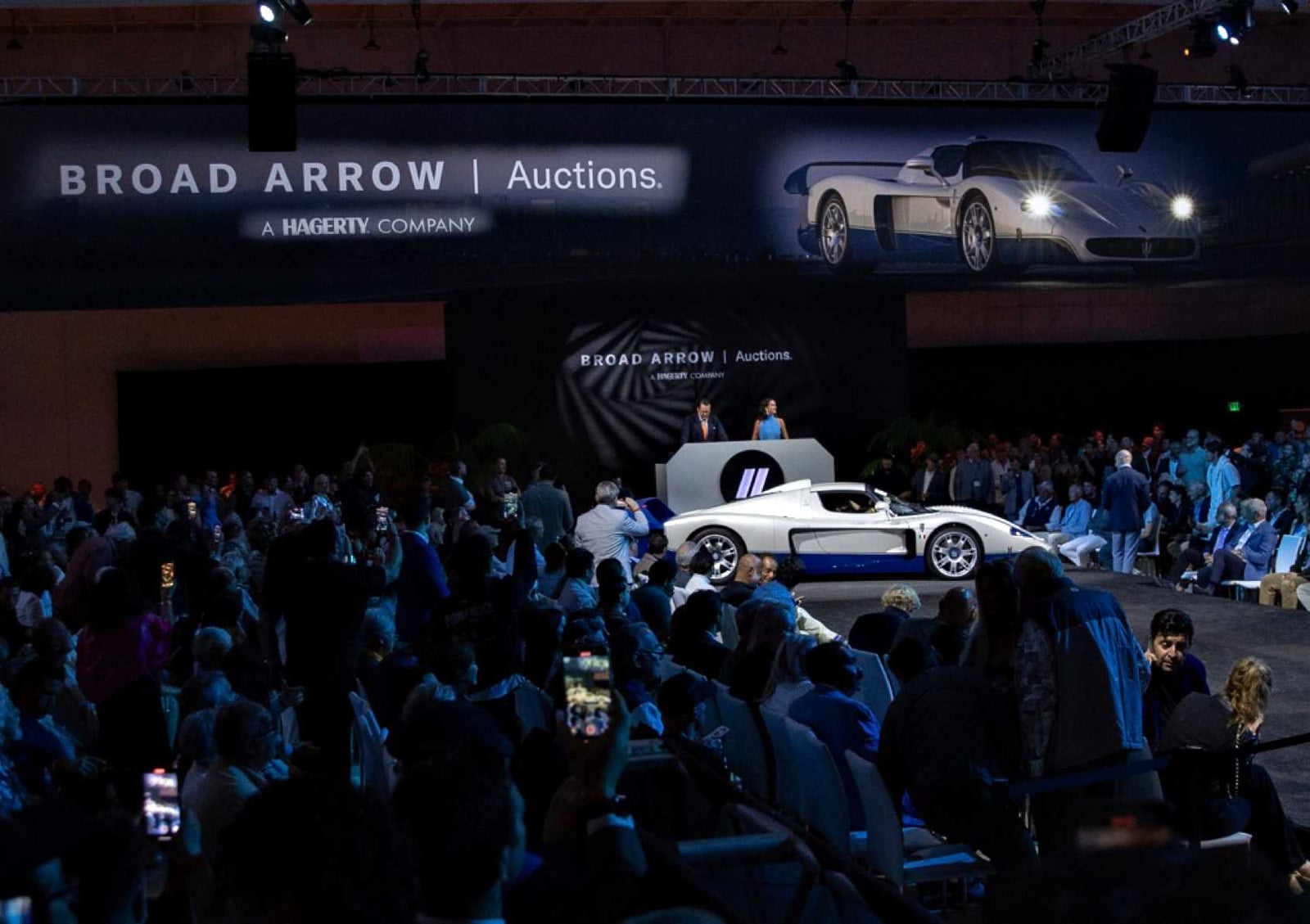 A white and blue sports car is displayed on stage at the Broad Arrow Jet Center Auction: Top 10 Results And Highlights event, surrounded by a seated audience and large automotive banners.