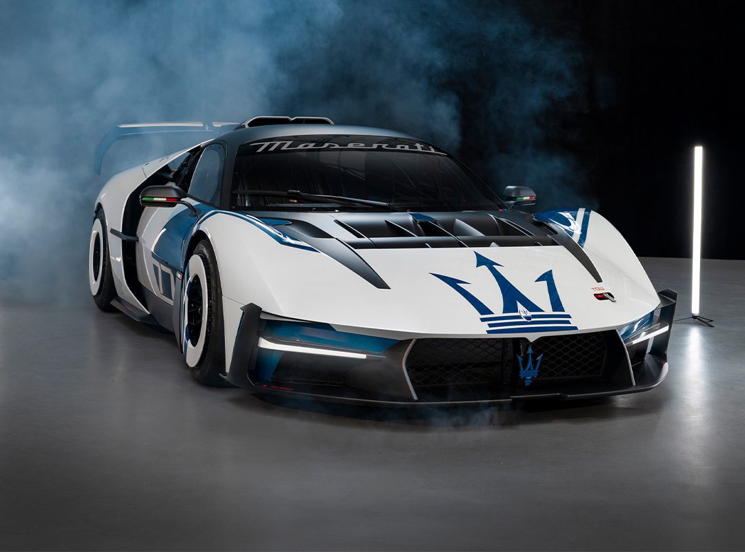 A white and black Maserati race car with blue accents and the Maserati trident logo on the hood, displayed indoors with smoke in the background.